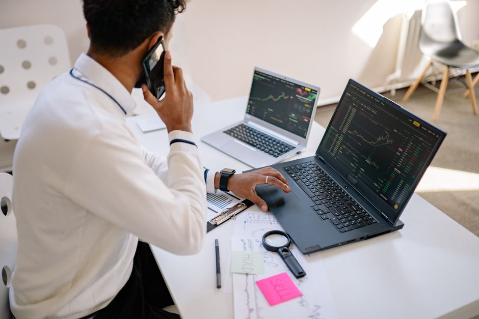 Profitable niche blog — Man holding phone, working on multiple laptops with financial charts at desk.